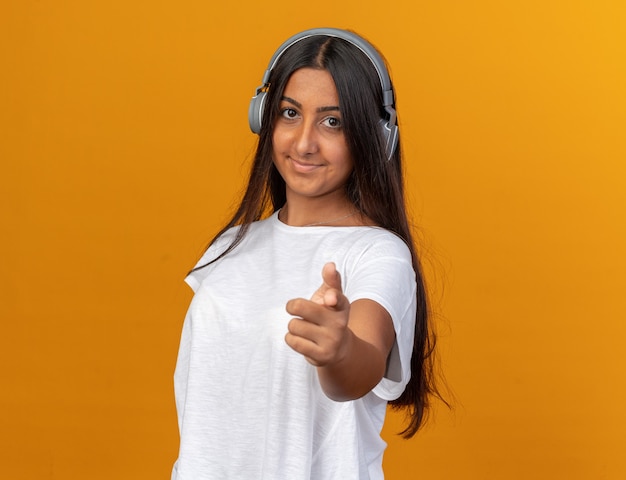 Chica joven en camiseta blanca con auriculares mirando a la cámara sonriendo alegremente apuntando con el dedo índice a la cámara de pie sobre naranja