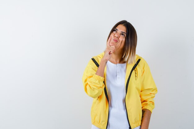 Chica joven en camiseta blanca, chaqueta amarilla apoyando la barbilla en la palma, pensando en algo y mirando pensativo, vista frontal.