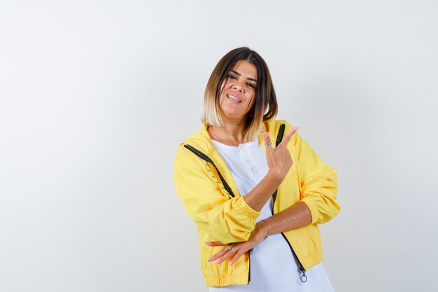 Chica joven en camiseta blanca, chaqueta amarilla apuntando a la derecha con el dedo índice y mirando alegre, vista frontal.