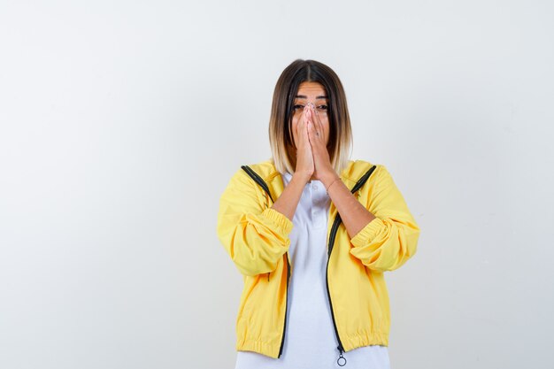 Chica joven en camiseta blanca, chaqueta amarilla juntando las manos delante de la boca y mirando emocionado, vista frontal.