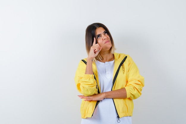 Chica joven en camiseta blanca, chaqueta amarilla levantando el dedo índice en gesto eureka y mirando sensible, vista frontal.