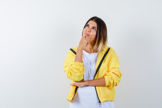 Chica joven en camiseta blanca, chaqueta amarilla de pie en pose de pensamiento, apoyando la barbilla en la mano y mirando pensativo, vista frontal.