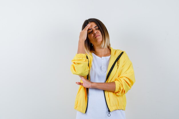 Chica joven con camiseta blanca, chaqueta amarilla poniendo la mano en la frente, con dolor de cabeza y luciendo agotada, vista frontal.