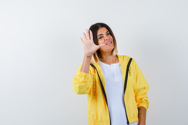 Chica joven en camiseta blanca, chaqueta amarilla que muestra la señal de stop y mirando alegre, vista frontal.