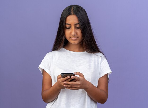 Foto gratuita chica joven en camiseta blanca escribiendo un mensaje de texto con smartphone parado sobre azul