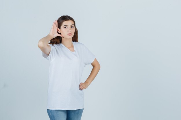 Chica joven en camiseta blanca escuchando una conversación privada y mirando curiosa, vista frontal.