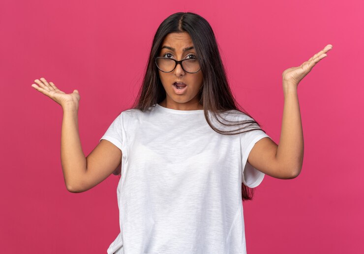 Foto gratuito chica joven en camiseta blanca con gafas mirando a la cámara confundida y sorprendida con los brazos levantados sobre rosa