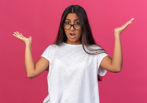 Chica joven en camiseta blanca con gafas mirando a la cámara confundida y sorprendida con los brazos levantados sobre rosa