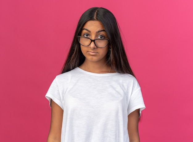 Chica joven en camiseta blanca con gafas mirando a la cámara con expresión seria y segura de pie sobre rosa