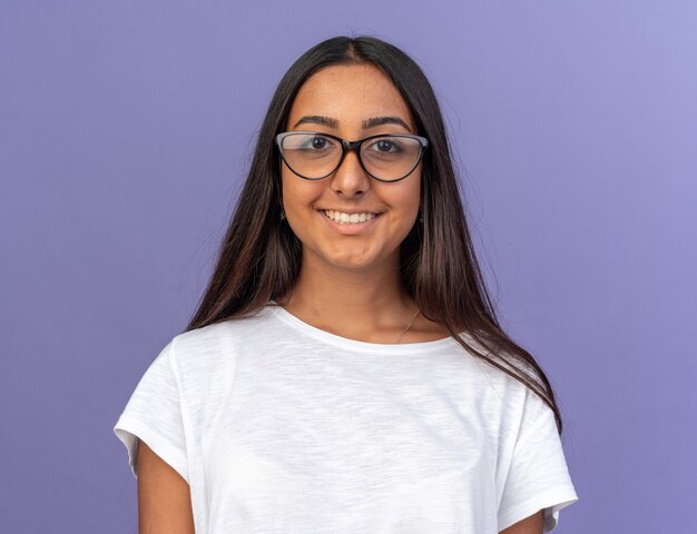Chica joven en camiseta blanca con gafas mirando a la cámara con una sonrisa en la cara feliz de pie sobre azul