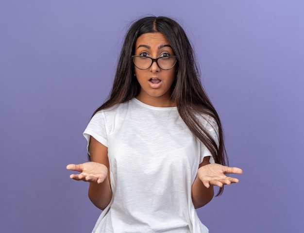 Chica joven en camiseta blanca con gafas mirando a la cámara sorprendido y asombrado de pie sobre fondo azul.