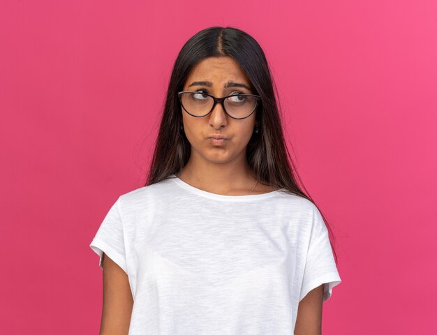 Chica joven en camiseta blanca con gafas mirando a un lado con expresión triste frunciendo los labios sobre rosa