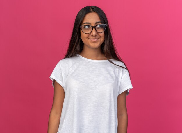 Chica joven en camiseta blanca con gafas mirando a un lado con una sonrisa en la cara feliz de pie sobre rosa