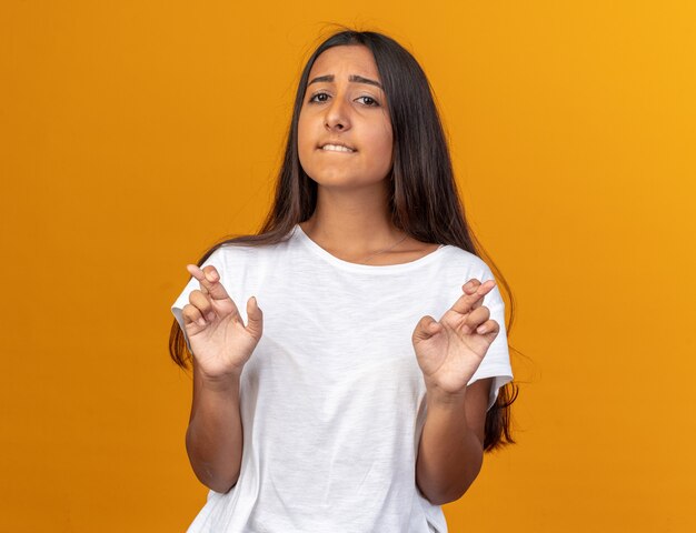 Chica joven en camiseta blanca haciendo deseo deseable cruzar los dedos con expresión de esperanza de pie sobre fondo naranja