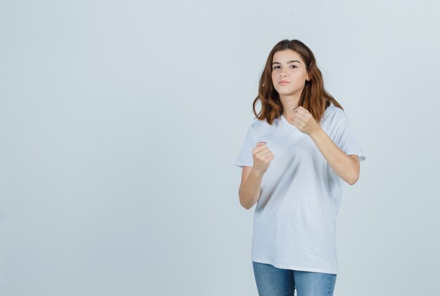 Chica joven en camiseta blanca, jeans de pie en pose de lucha y mirando confiado, vista frontal.