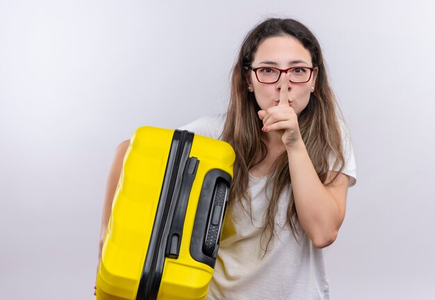 Chica joven en camiseta blanca con maleta de viaje haciendo gesto de silencio con el dedo en los labios