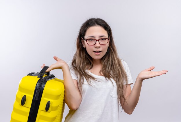 Chica joven en camiseta blanca con maleta de viaje mirando incierto y confundido extendiendo las palmas