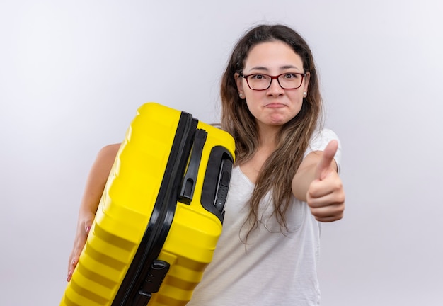 Chica joven en camiseta blanca con maleta de viaje sonriendo mostrando los pulgares para arriba