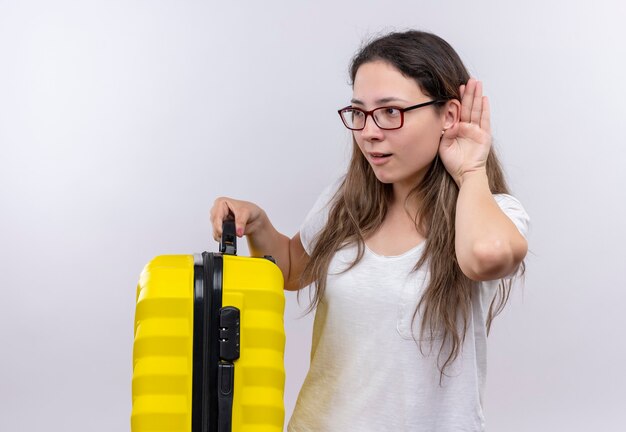 Chica joven en camiseta blanca con maleta de viaje sosteniendo la mano cerca del oído tratando de escuchar la conversación de alguien