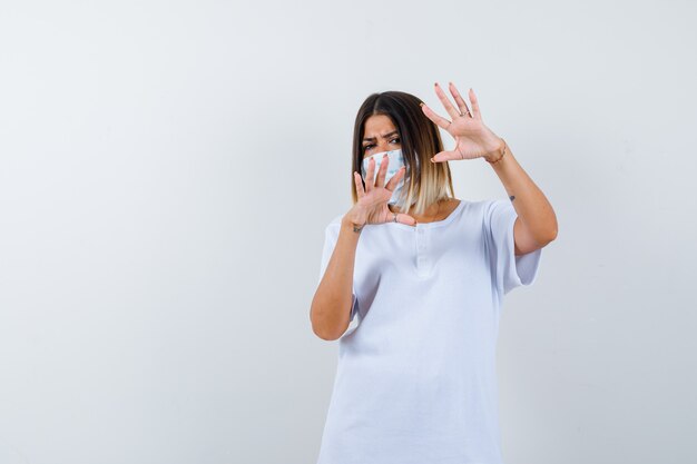 Chica joven en camiseta blanca, máscara mostrando gesto de restricción y mirando serio, vista frontal.