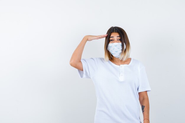 Chica joven en camiseta blanca y máscara mostrando gesto de saludo y mirando confiado, vista frontal.