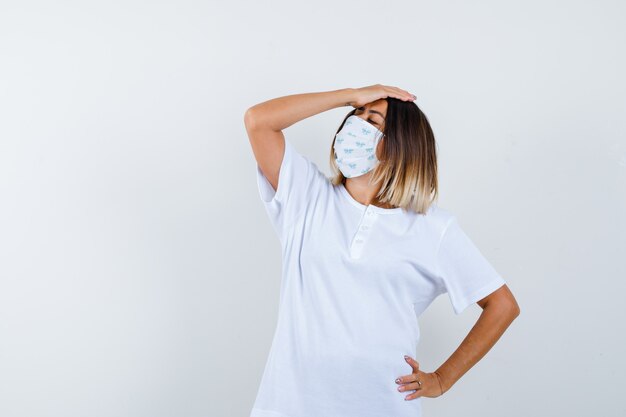 Chica joven en camiseta blanca y máscara poniendo una mano en la cabeza, otra mano en la cintura y mirando preocupado, vista frontal.