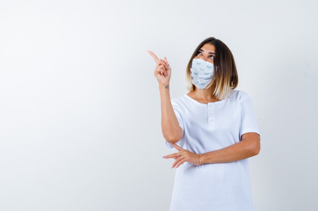 Chica joven en camiseta blanca y máscara sosteniendo una mano debajo del codo, apuntando hacia arriba con el dedo índice y mirando enfocado, vista frontal.