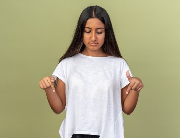 Chica joven en camiseta blanca mirando hacia abajo con la cara seria apuntando hacia abajo con los dedos índices de pie sobre verde