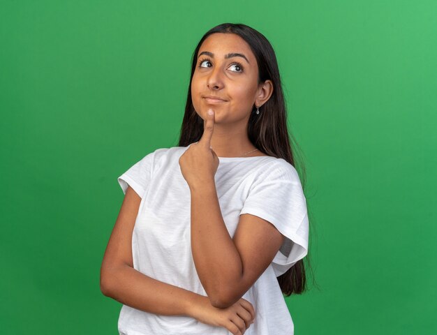 Chica joven en camiseta blanca mirando hacia arriba perplejo parado sobre fondo verde