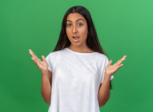 Chica joven en camiseta blanca mirando a la cámara asombrada y sorprendida con los brazos levantados de pie sobre fondo verde