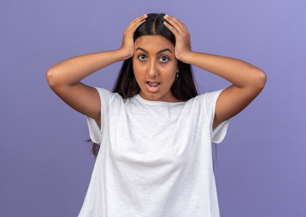 Chica joven en camiseta blanca mirando a la cámara asombrada y sorprendida con las manos en la cabeza de pie sobre fondo azul.