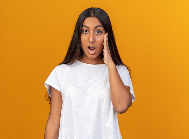 Chica joven en camiseta blanca mirando a la cámara asombrada y sorprendida de pie sobre fondo naranja