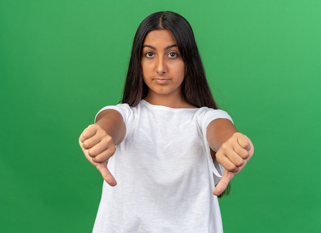 Chica joven en camiseta blanca mirando a cámara con cara seria mostrando los pulgares hacia abajo