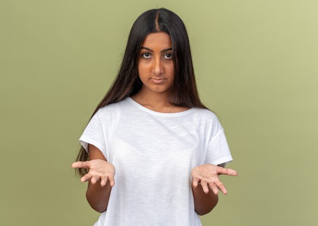 Chica joven en camiseta blanca mirando a la cámara confundida levantando los brazos con disgusto e indignación de pie sobre verde