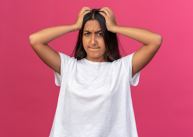 Foto gratuita chica joven en camiseta blanca mirando a la cámara confundida y muy ansiosa con las manos en la cabeza sobre rosa