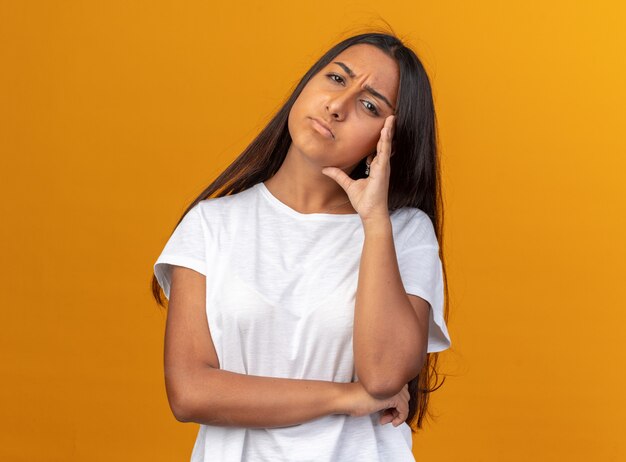 Chica joven en camiseta blanca mirando a la cámara disgustado cansado y aburrido de pie sobre fondo naranja
