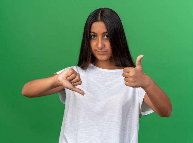 Chica joven en camiseta blanca mirando a la cámara disgustado mostrando los pulgares hacia arriba y hacia abajo de pie sobre fondo verde