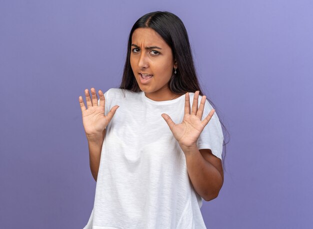 Chica joven en camiseta blanca mirando a la cámara disgustado sosteniendo las manos como diciendo no te acerques