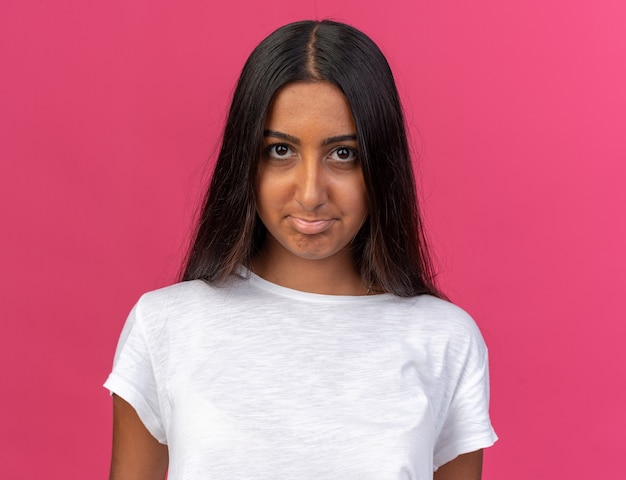 Foto gratuita chica joven en camiseta blanca mirando a la cámara con expresión triste frunciendo los labios sobre fondo rosa