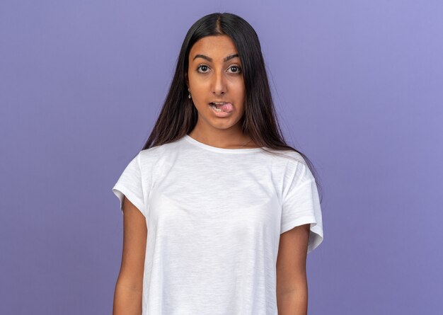 Chica joven en camiseta blanca mirando a la cámara feliz y alegre sacando la lengua de pie sobre fondo azul.