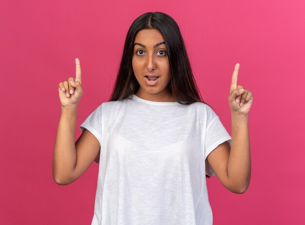 Chica joven en camiseta blanca mirando a cámara feliz y emocionado mostrando los dedos índices teniendo nueva idea de pie sobre fondo rosa