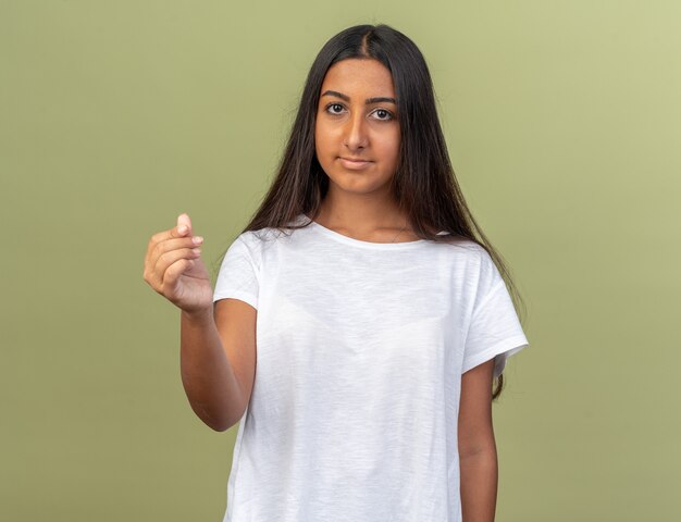 Chica joven en camiseta blanca mirando a la cámara haciendo gesto de dinero frotándose los dedos