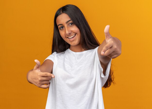 Chica joven en camiseta blanca mirando a la cámara sonriendo alegremente apuntando con índice figners en la cámara de pie sobre naranja