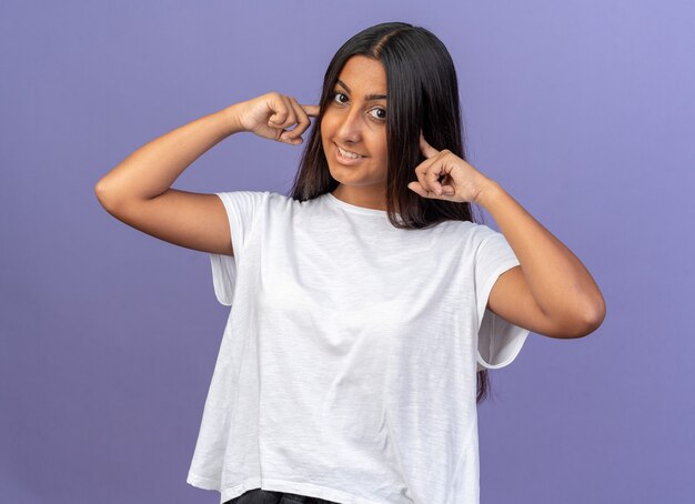 Chica joven en camiseta blanca mirando a la cámara sonriendo alegremente cerrando las orejas con los dedos tanding sobre fondo azul.