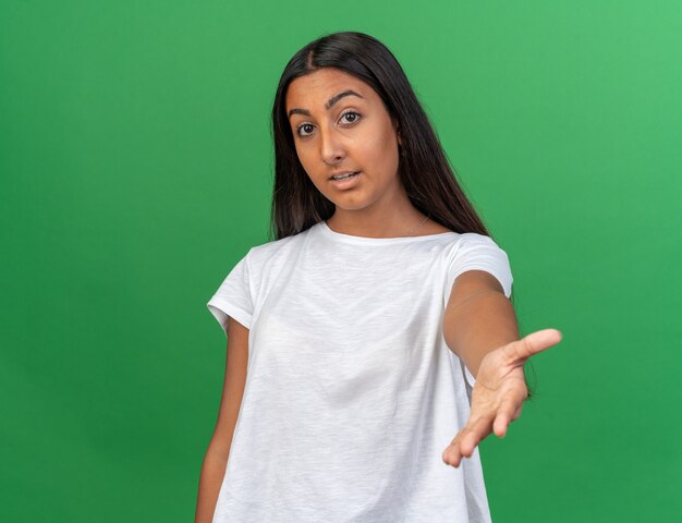 Chica joven en camiseta blanca mirando a la cámara sonriendo amable haciendo ven aquí gesto de pie sobre fondo verde