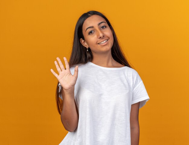 Chica joven en camiseta blanca mirando a la cámara sonriendo amistosamente saludando con la mano