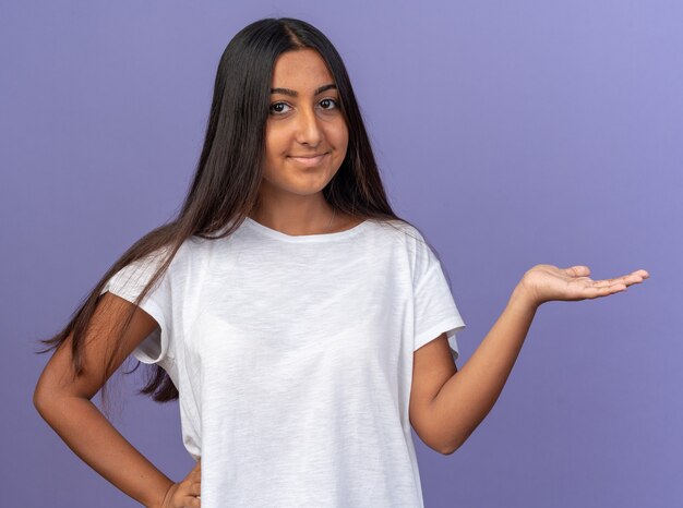 Chica joven en camiseta blanca mirando a la cámara sonriendo confiado presentando copia espacio con el brazo de la mano de pie sobre fondo azul.