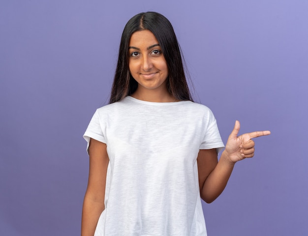 Chica joven en camiseta blanca mirando a la cámara con una sonrisa en la cara feliz apuntando con el dedo índice hacia el lado de pie sobre azul