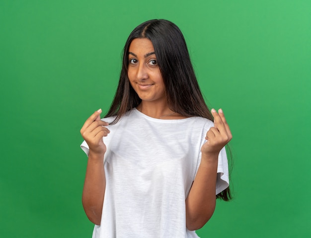Chica joven en camiseta blanca mirando a la cámara con una sonrisa en la cara haciendo gesto de dinero frotando los dedos