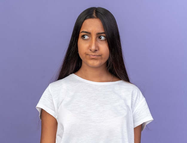Chica joven en camiseta blanca mirando a un lado con cara seria de pie sobre azul